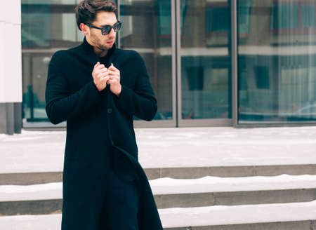 Portrait of young trendy man wearing a black coat with a raised collar in sunglasses. Streetstyle. On the background a business building. Frozen on the street. Fashion.の写真素材