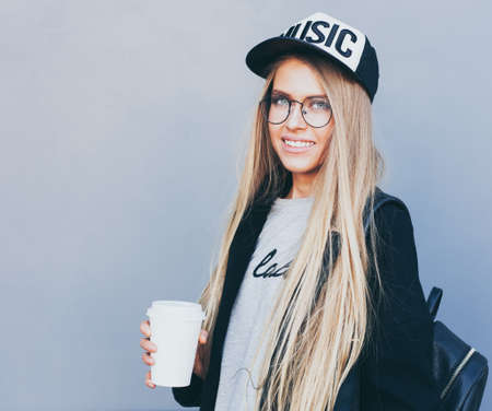 Closeup portrait of blonde girl with long haircut. Hipster, insagram style. She wears black trendy dress, glasses, backpack, baseball cap. She is looking to the camera. Outdoor.の写真素材