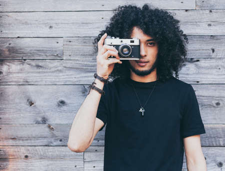 African american man with typical afro hair takes a picture of a vintage camera against a background of old boardsの写真素材