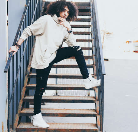 Young fashionably dressed man with an afro hairstyle and a beard in a cool outfit posing on a metal staircase. youth fashion Streetstyle. Youth fashion.の写真素材