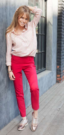 Portrait of a young woman dressed in a blouse, red Chino Trouser, and gold brogues. Posing next to a gray wall. Fashion. Streetstyle.の写真素材