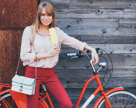 Beautiful woman eating ice cream. Dressed a red Chino Trouser and gold sneakers, fashionable stylish clothes. Outdoor. Red vintage bike.の写真素材