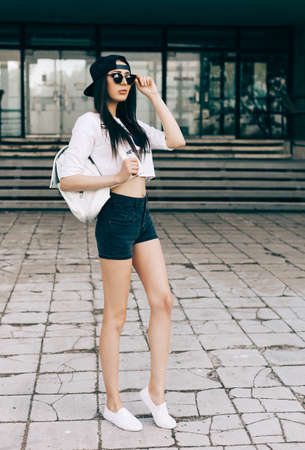A street photo of a young beautiful female wearing white t-shirt and black jeans shorts standing on...