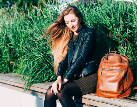 Beautiful blond long haired hipster young woman with trendy leather sitting on park bench on summer evening. Outdoor.の写真素材