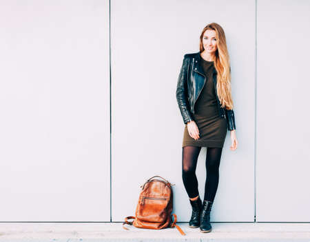 Beautiful young woman with long hair posing with fashionable backpack near the gray wallの写真素材