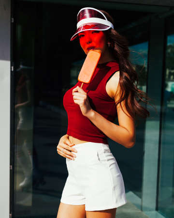 Sunny portrait of an Long-haired Asian girl in a trendy summer outfit, fashionable red Brim Visor Hat is enjoyed by ice cream.の写真素材
