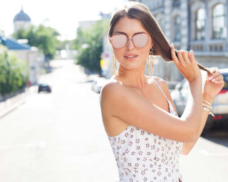 Portrait of a girl in a light white dress and sunglasses makes a hairstyle on a hot day on the European streetの写真素材