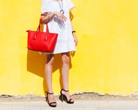 Fashionista shopaholic posing in a light summer dress and a red big handbag on a yellow wall background. Part of body.の写真素材