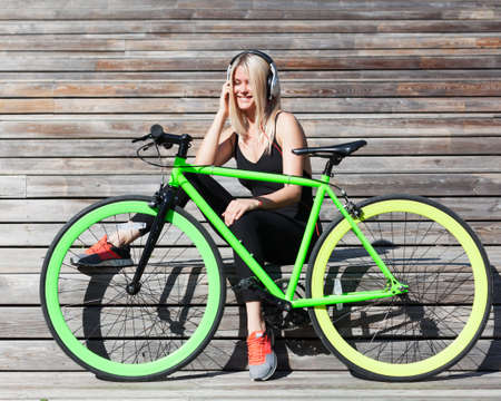 Fitness princess with long blond hair girl in black sexy outfit and sneakers resting on wooden boards with fashionable fix bike listening to music on headphones on a sunny afternoonの写真素材