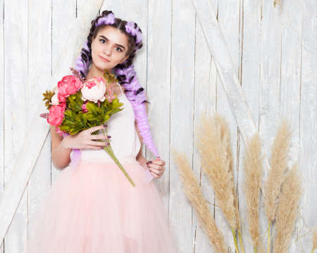 Smiling beautiful young girl with colored braids in a light outfit holding flowers in her hand and posing on a wooden background, indoor.の写真素材