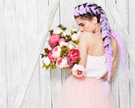 A young girl in a light outfit stands her back with flowers. Hairstyle with colored braids. Indoor.