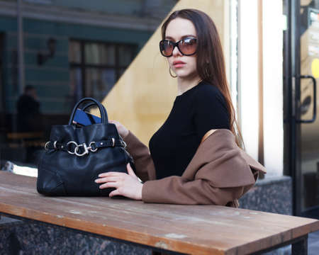 A young woman posing in a trendy street cafe on a street in a European city, a coat, a black dress, sunglasses and a large black handbag. Fashion style. Outdoor. Fashion bloggerの写真素材