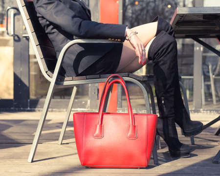 A young blond woman fashionable blogger sits in a street cafe in the European city in a black trendy suit, high heels and a big red handbag. Part of the body.の写真素材