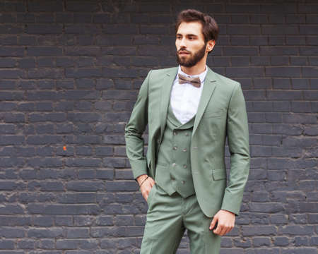 Young businessman with a beard in a fashionable green suit posing against a black brick wallの写真素材