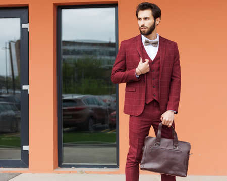 A young businessman with a beard in a trendy burgundy suit and a trendy handbag poses next to the shops in the city center. The trader.の写真素材