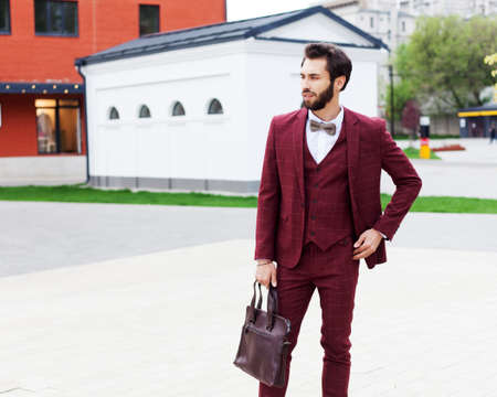 A young businessman with a beard in a trendy burgundy suit and a trendy handbag poses next to the shops in the center of the European city. The trader.の写真素材