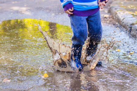 A little boy is jumping in a puddle. A boy in rubber boots. Happy childhood. Puddles after the rain. Warm summer evening. Kids gamesの写真素材