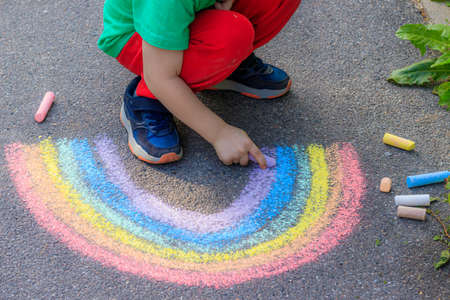 A boy draws a rainbow on the asphalt with crayons. A child draws on the asphalt. Children's drawing. Walking in the city with a child. Summer in the city. Crayons for drawing. Article about childhood.の写真素材