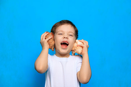 A boy listens to a shell on a blue background. Dreams of the sea. Article about vacation. Childhood. Baby and shell. Blue background. Copy spaceの写真素材