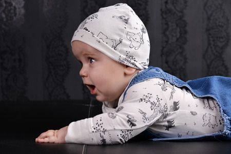 Cute baby boy in a black and white suit and rifles crawling on black sofa with black wallpaper in...