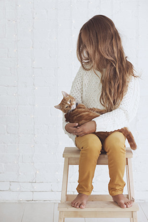 Cute teenage girl 8-9 years old wearing knit trendy winter clothes holding her ginger cat posing over white brick wallの写真素材
