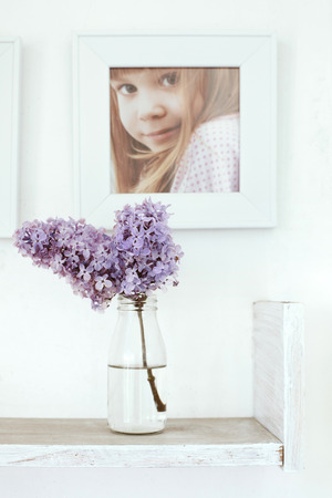 Small bouquet of lilac in glass bottle on white wooden shelf with photo frame on it as detail of interiorの写真素材