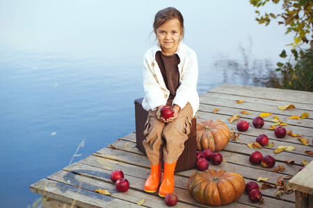 Cute little girl sitting on a vintage suitcase near the lake in warm autumn day. Halloween pumpkins, apples and fallen leaves beside.の写真素材