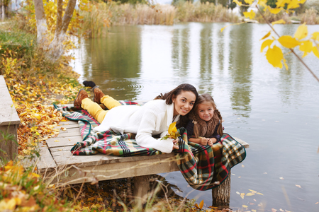 Young mother with her little daughter walking in fall park near pond one autumn dayの写真素材