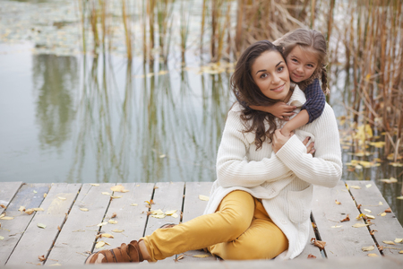 Young mother with her little daughter walking in fall park near pond one autumn dayの写真素材