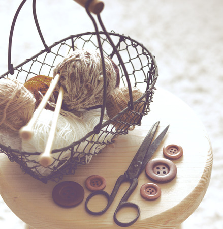 Vintage knitting needles, scissors and yarn inside old wire basket on wooden stool, still life photo with soft focusの写真素材