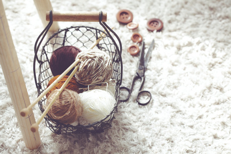 Vintage knitting needles, scissors and yarn inside old wire basket on wooden stool, still life photo...