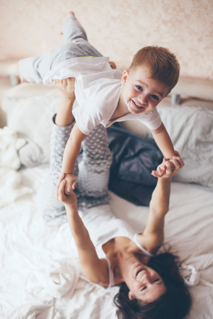 Young mother with her 2 years old little son dressed in pajamas are relaxing and playing in the bed at the weekend together, lazy morning, warm and cozy scene. Pastel colors, selective focus.の写真素材