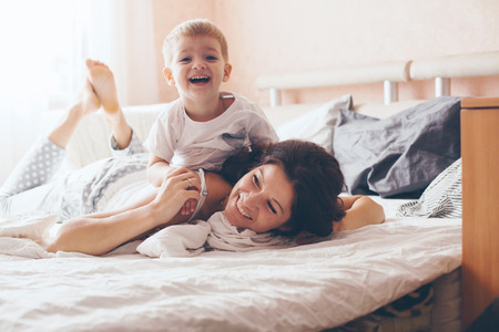 Young mother with her 2 years old little son dressed in pajamas are relaxing and playing in the bed at the weekend together, lazy morning, warm and cozy scene. Pastel colors, selective focus.の写真素材