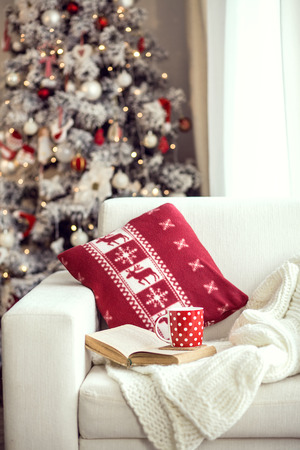 Opened book and a cup of tee on the cozy chair with warm blanket and cushion on it near Christmas treeの写真素材