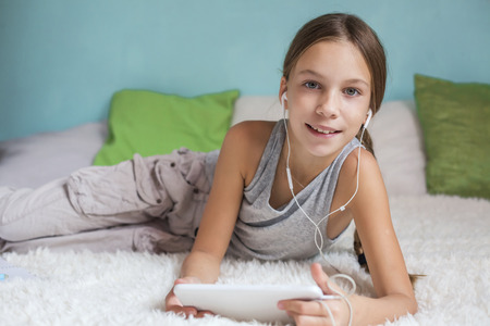 Pre teen girl is relaxing in the bed and listening to music with earphones on the tablet at homeの写真素材