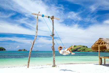 Woman in white dress and hat swinging at tropical beach, sunny day, good weatherの写真素材