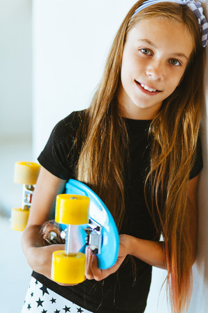 Pre teen girl wearing cool fashion clothing posing with colorful skateboard against white wallの写真素材
