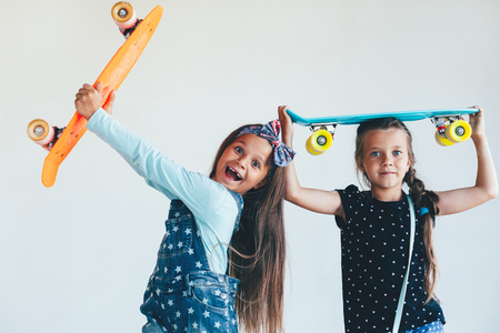 Two 7 years old children wearing cool fashion jeans clothing posing with colorful skateboard against white wall, urban styleの写真素材