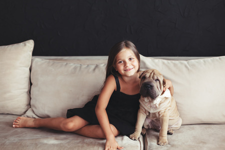 Home portrait of cute child hugging with puppy of Chinese Shar Pei dog on the sofa against black wallの写真素材