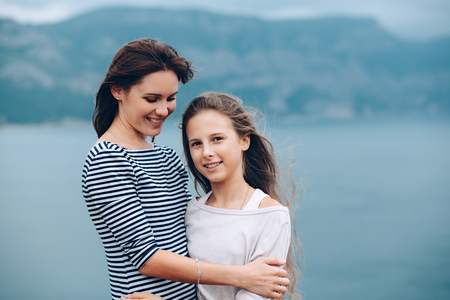 Mom and her teenage daughter hugging and smiling together over blue sea viewの写真素材