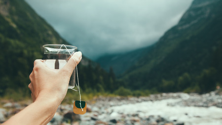 Closeup photo of mug with tea bag in traveler's hand over out of focus mountains view, tourizm in cold seasonの写真素材