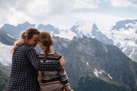 Mom with daughter relaxing in the rustick wooden terrace on mountain, alpine view, snow on hills. Dombay, Karachay-Cherkessia, Caucasus, Russia.の写真素材