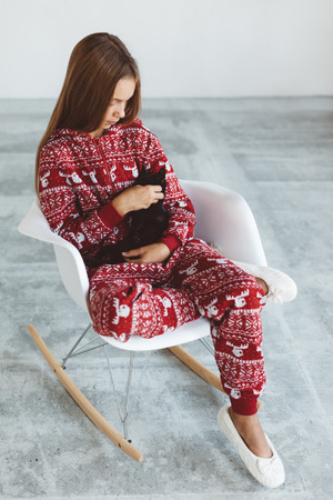 10 years old child dressed in warm Christmas pajamas sitting with black kitten on modern rocking chair in concrete scandinavian interior. Lazy winter morning, comfortable scene.の写真素材