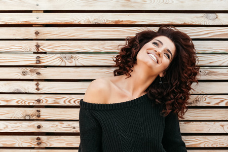 Beautiful friendly girl with curly hair wearing black sweater posing against wooden wall, looking up...