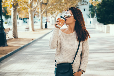 Young woman wearing woolen sweater walking in the autumn city street and drinking take away coffee in paper cup. Breakfast on the go.の写真素材