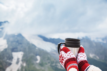 Closeup photo of thermos mug with tea in traveler's hand over out of focus mountains view with snow, tourizm in cold seasonの写真素材