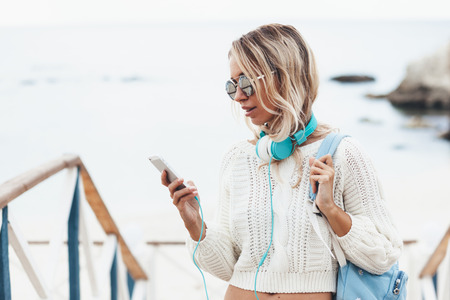 Young woman with backpack wearing white sweater relaxing and listening music at the beach, autumn weekend on the sea shoreの写真素材