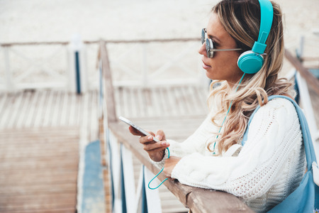 Young woman with backpack wearing white sweater relaxing and listening music at the beach, autumn weekend on the sea shoreの写真素材