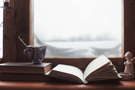 Warm and comfy winter concept. Book, cup of tea and candlestick on wooden window sill in old house. Reading and relaxing in cold snowy weather at home. Quiet silent homely scene.の写真素材
