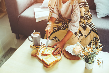 Unrecognizable girl wrapped in blanket relaxing with cocoa and breakfast on a couch in living room....